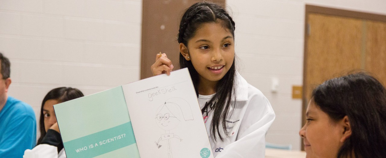 A young child in a lab coat shows off her drawing of what a scientist looks like.